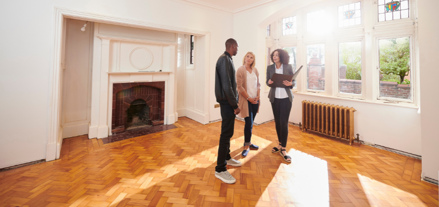 A real estate agent showing an empty, well-lit living space with wooden flooring and a fireplace to two potential buyers.